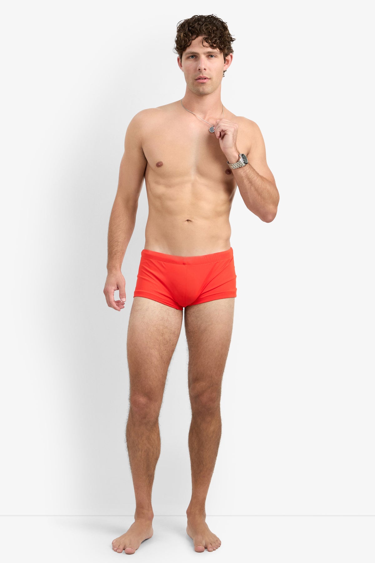 A barefoot man with curly hair stands against a plain white background, wearing the bright red Montego Bay Swim Trunk. He is shirtless, holding a necklace, and looking directly at the camera.