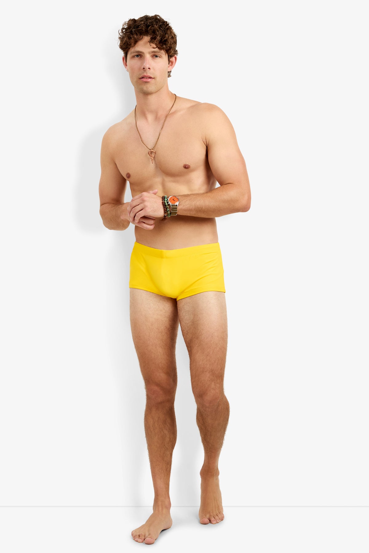 A man with curly brown hair stands barefoot against a white background, wearing the Montego Bay Swim Trunk. He is shirtless, accessorized with a necklace and colorful bracelets, facing forward with a neutral expression.