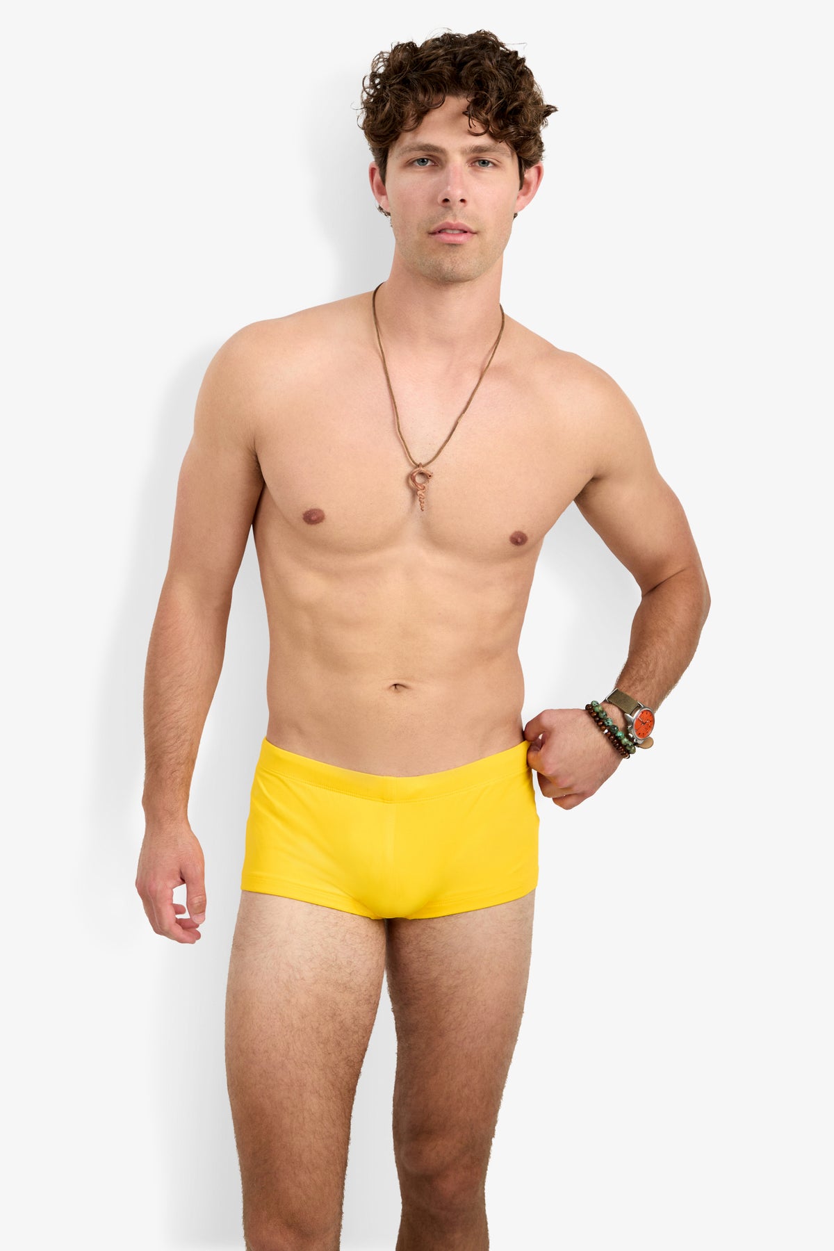 A shirtless man with curly brown hair poses against a white background, wearing the Montego Bay Swim Trunk in bright yellow, along with a necklace and bracelets on his left wrist.