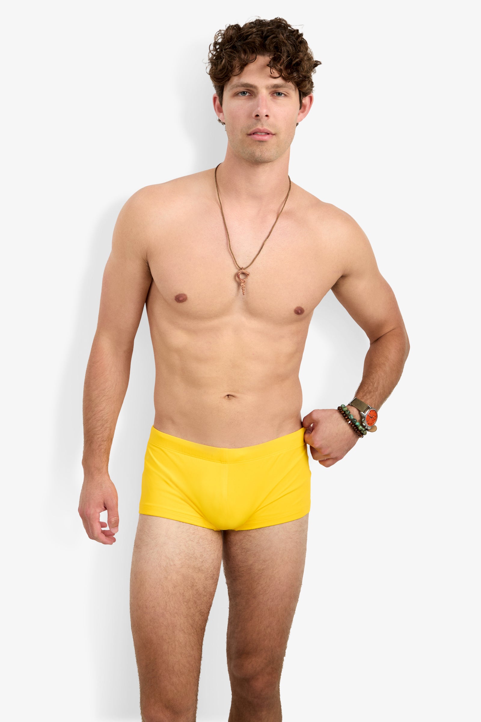 A man with curly brown hair, wearing a necklace and bracelets, poses against a plain background in bright yellow Montego Bay Swim Trunks.