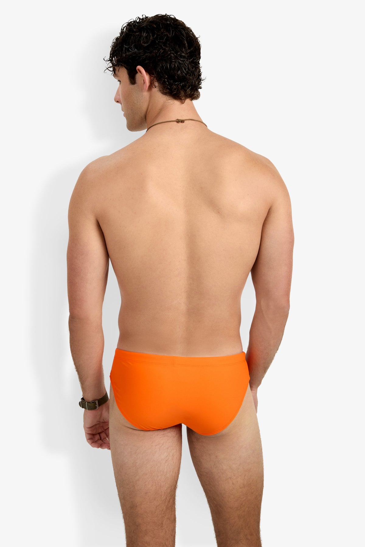 A man with short curly hair stands with his back to the camera in a Palm Beach Swim Brief, wearing a watch on his left wrist and a necklace. The background is plain white.
