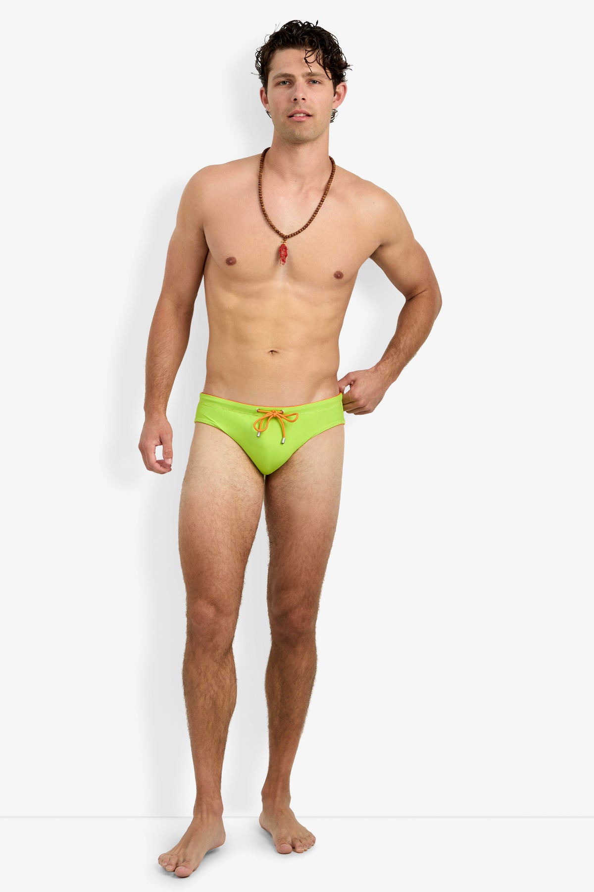 A man with short, wavy brown hair and a relaxed expression stands barefoot against a white background, wearing the Palm Beach Swim Brief in bright green with an orange drawstring and a beaded necklace.