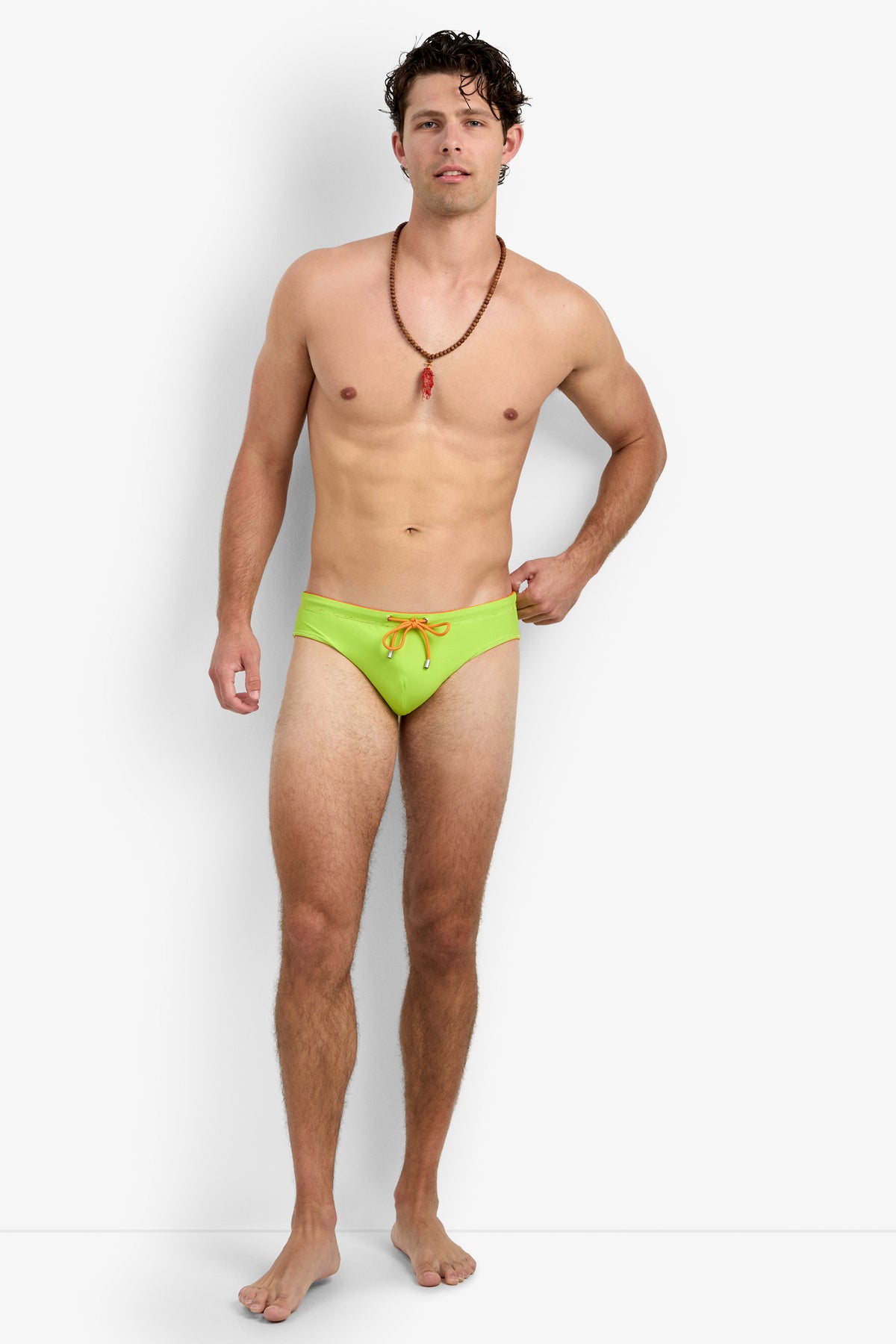 A man with short curly hair and a beaded necklace stands barefoot against a plain white background, wearing bright green Palm Beach Swim Briefs.
