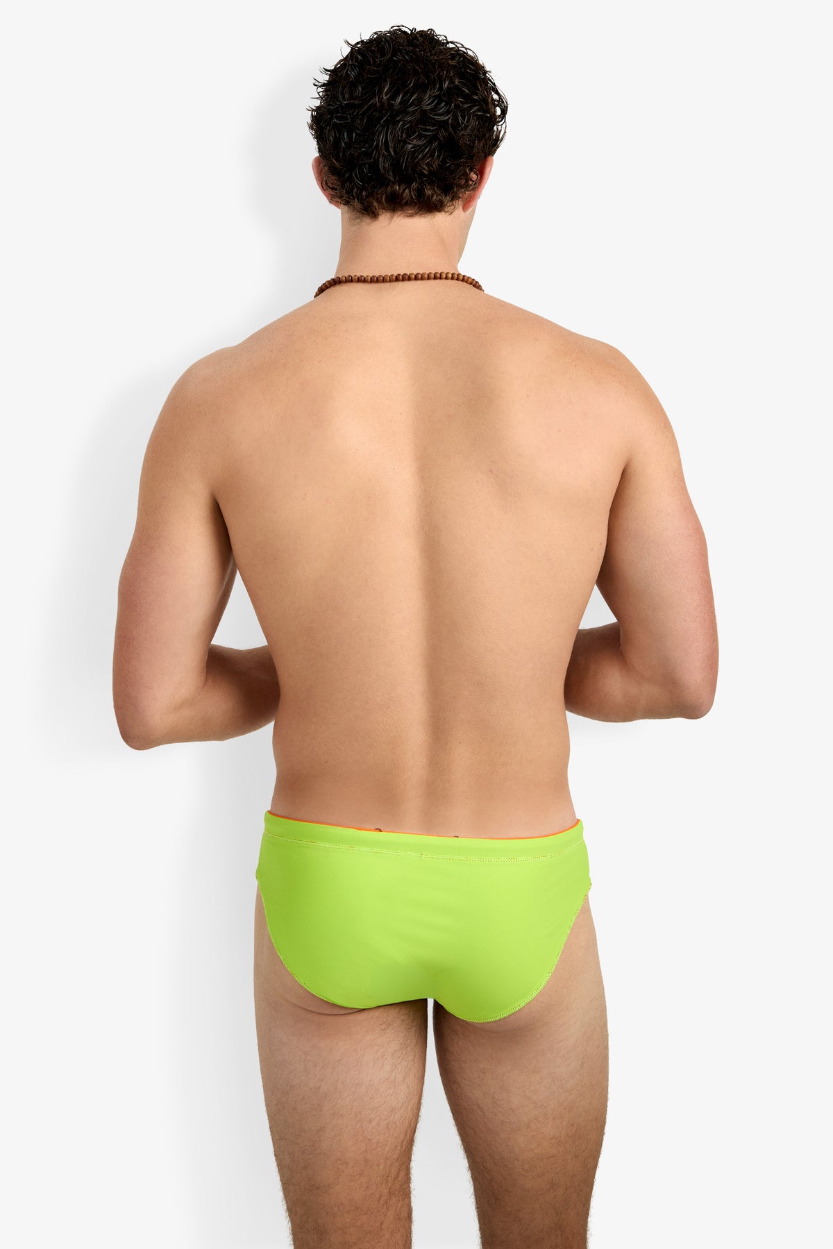 A man is shown from the back wearing the Palm Beach Swim Brief in bright green and a beaded necklace, standing against a plain white background.
