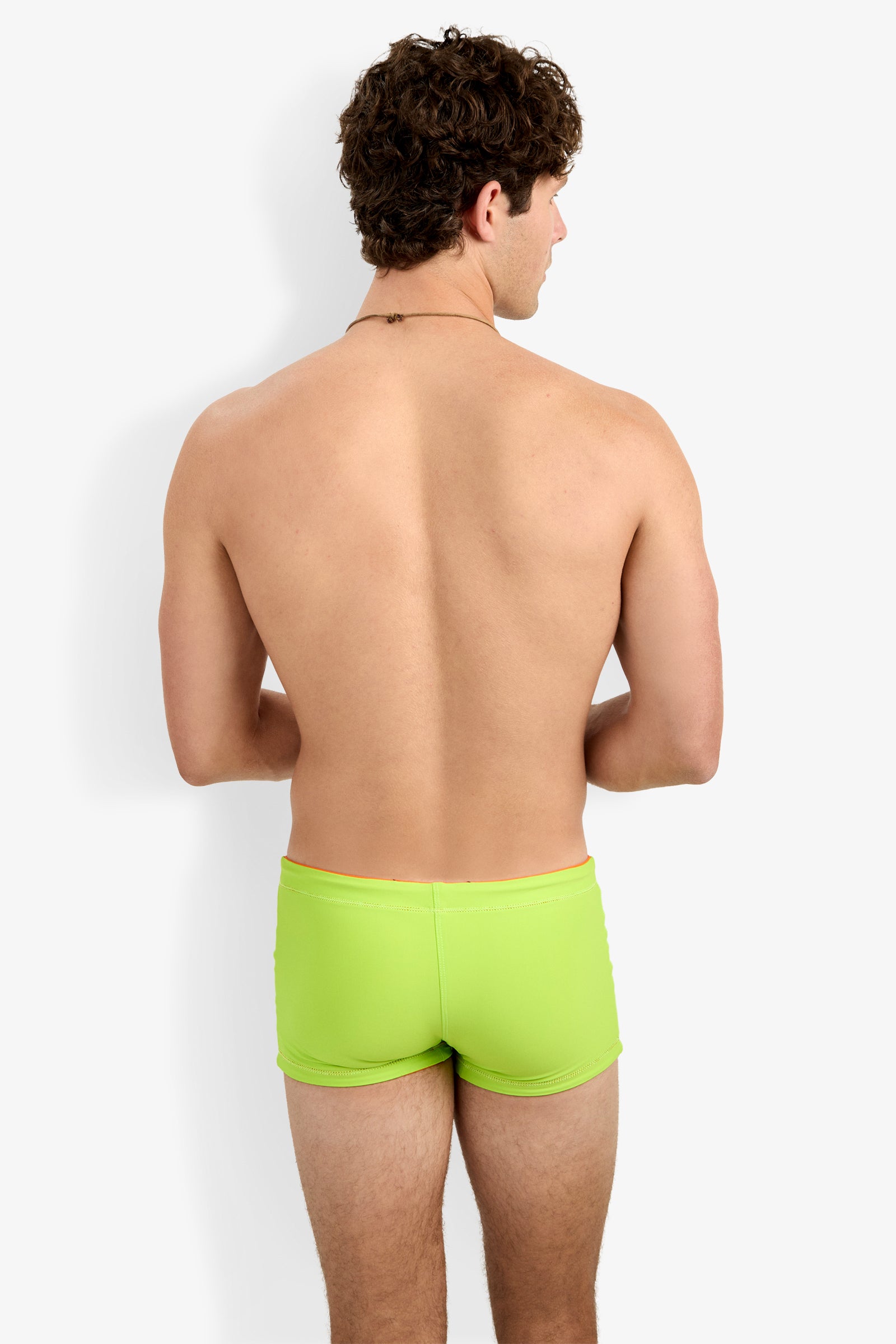 A shirtless man with curly brown hair faces away, wearing Seaside Beach Swim Trunk in bright green against a plain white background.