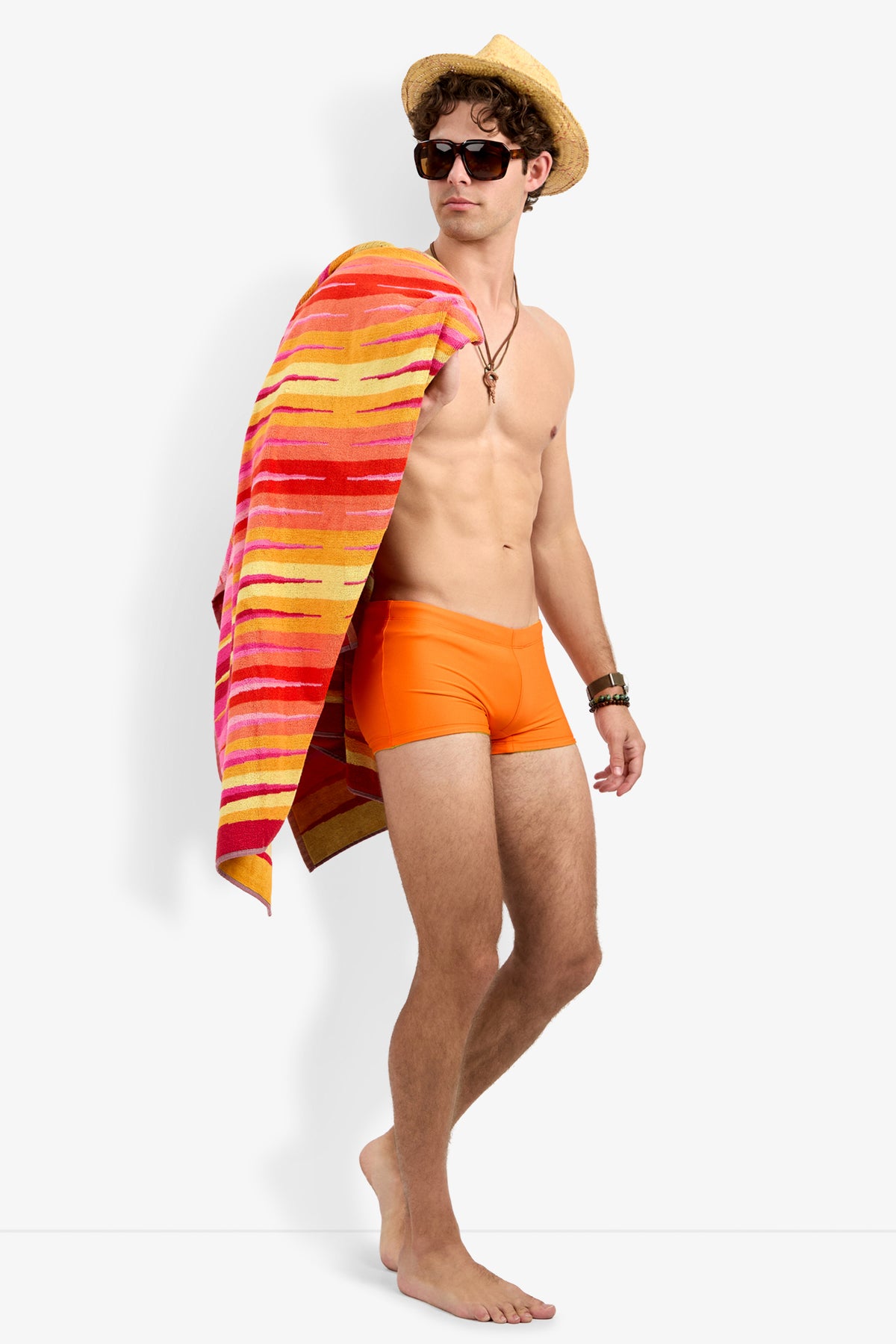 A man wearing Seaside Beach Swim Trunk, sunglasses, and a straw hat stands barefoot with a colorful striped towel over his shoulder, looking to the side.