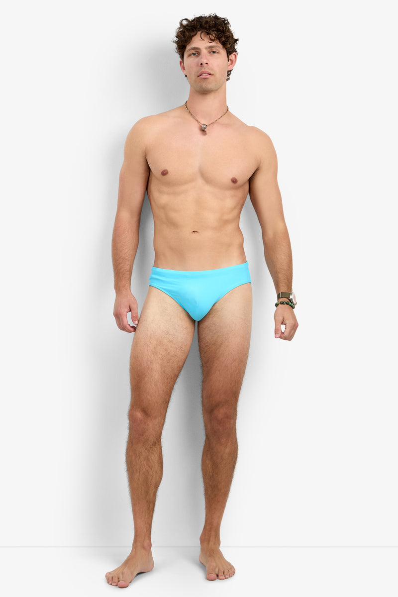A man stands barefoot against a plain white background, wearing the Palm Beach Swim Brief. He faces the camera with a neutral expression, also accessorized with a necklace and wristwatch.