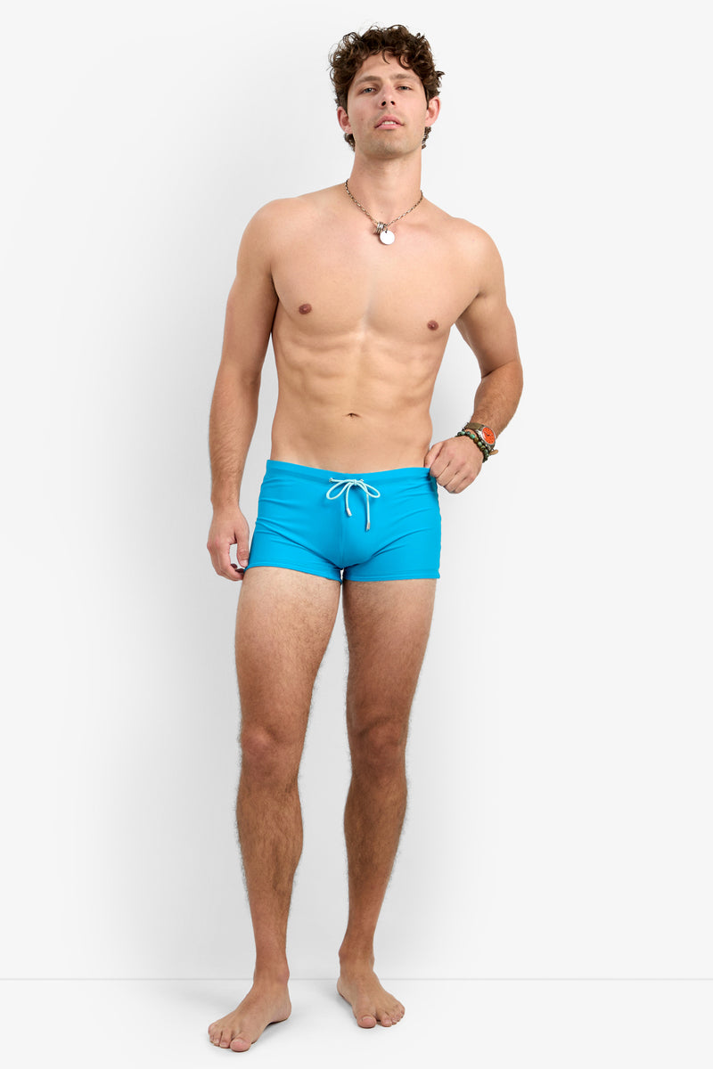A man with curly brown hair and a relaxed expression stands barefoot against a white background, wearing the Seaside Beach Swim Trunk in bright blue with a white drawstring and a necklace.