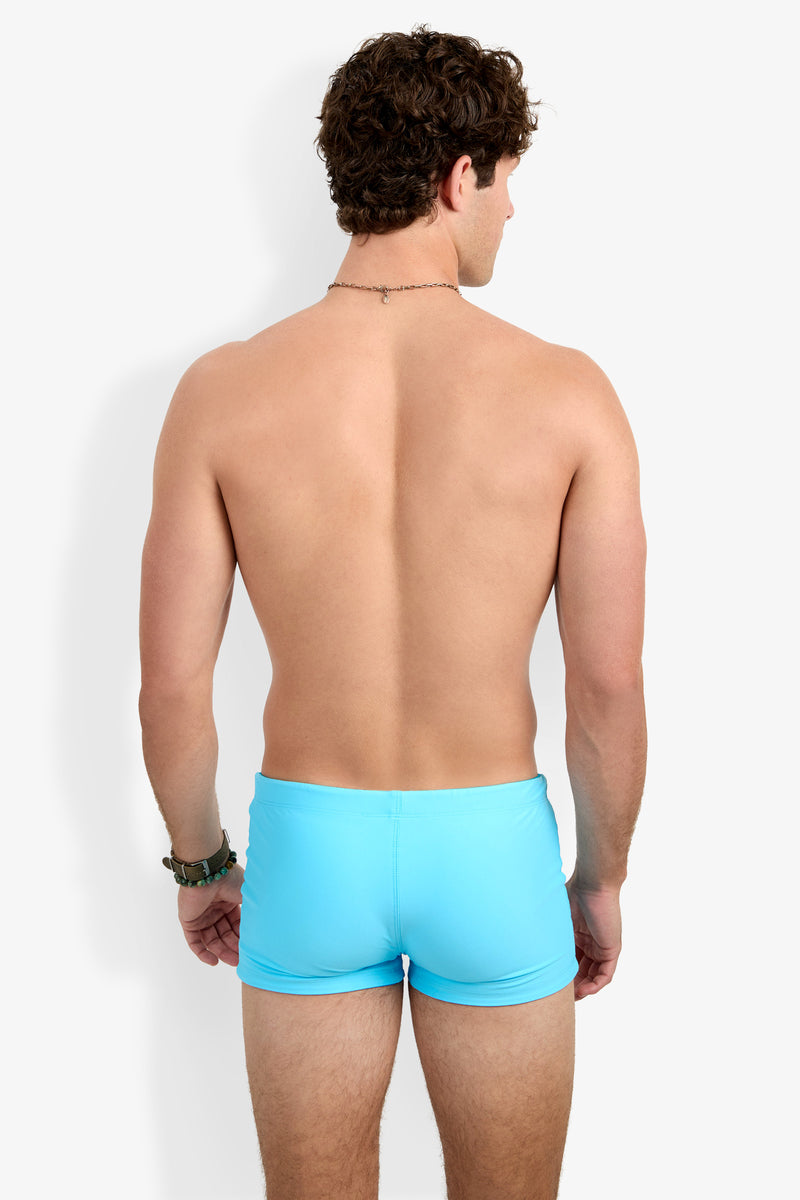 A shirtless man with short curly hair, beaded necklace, and bracelets faces away from the camera, modeling the Seaside Beach Swim Trunk in light blue against a plain white background.