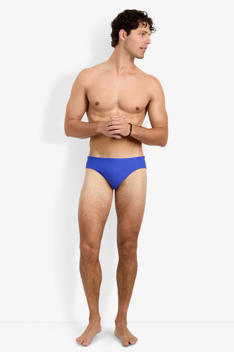 A shirtless man wearing the Palm Beach Swim Brief in blue stands barefoot against a plain white background, facing slightly to the side with his hands loosely clasped in front of him.