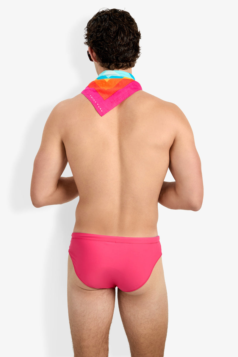 A man with short, curly dark hair stands with his back to the camera against a plain white background, wearing the bright pink Palm Beach Swim Brief and a colorful bandana around his neck.