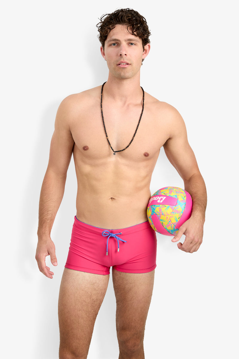A man wearing Seaside Beach Swim Trunk stands holding a colorful volleyball under one arm against a plain white background.