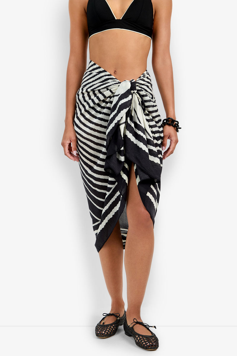 A person wearing a black bikini top, the Pareo Swim Cover-Up in black-and-white stripes tied at the waist, black shoes, and a black bracelet stands against a plain white background.