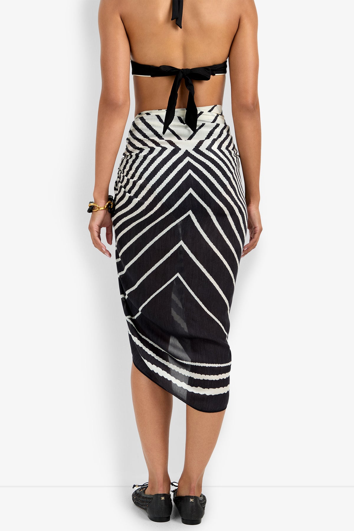 A person is seen from the back wearing a black halter top and the Pareo Swim Cover-Up, which has a black-and-white geometric chevron pattern and an asymmetrical design, paired with black shoes.