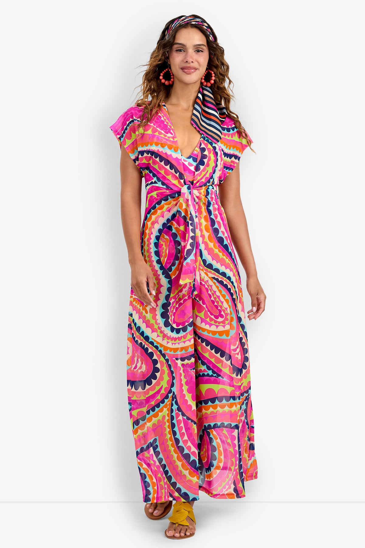 A woman models the Cover-Up Maxi Dress, featuring a vibrant multicolored wavy pattern in pink, orange, blue, and yellow. She completes the look with a matching headband, red earrings, and yellow sandals against a white background.