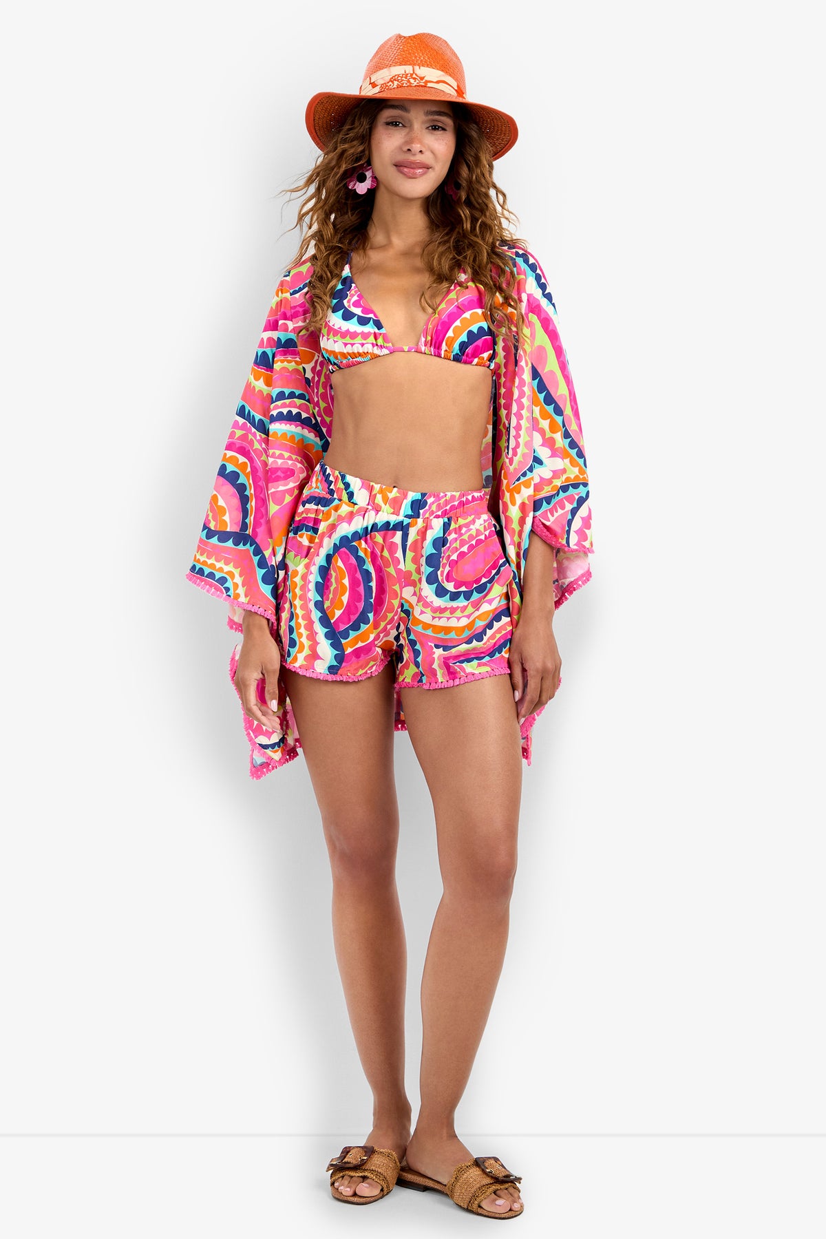 Woman wearing a Kimono Swim Cover-up over a colorful wavy-patterned swimsuit set with shorts, paired with a wide-brimmed orange hat, pink earrings, and brown sandals, standing and smiling.