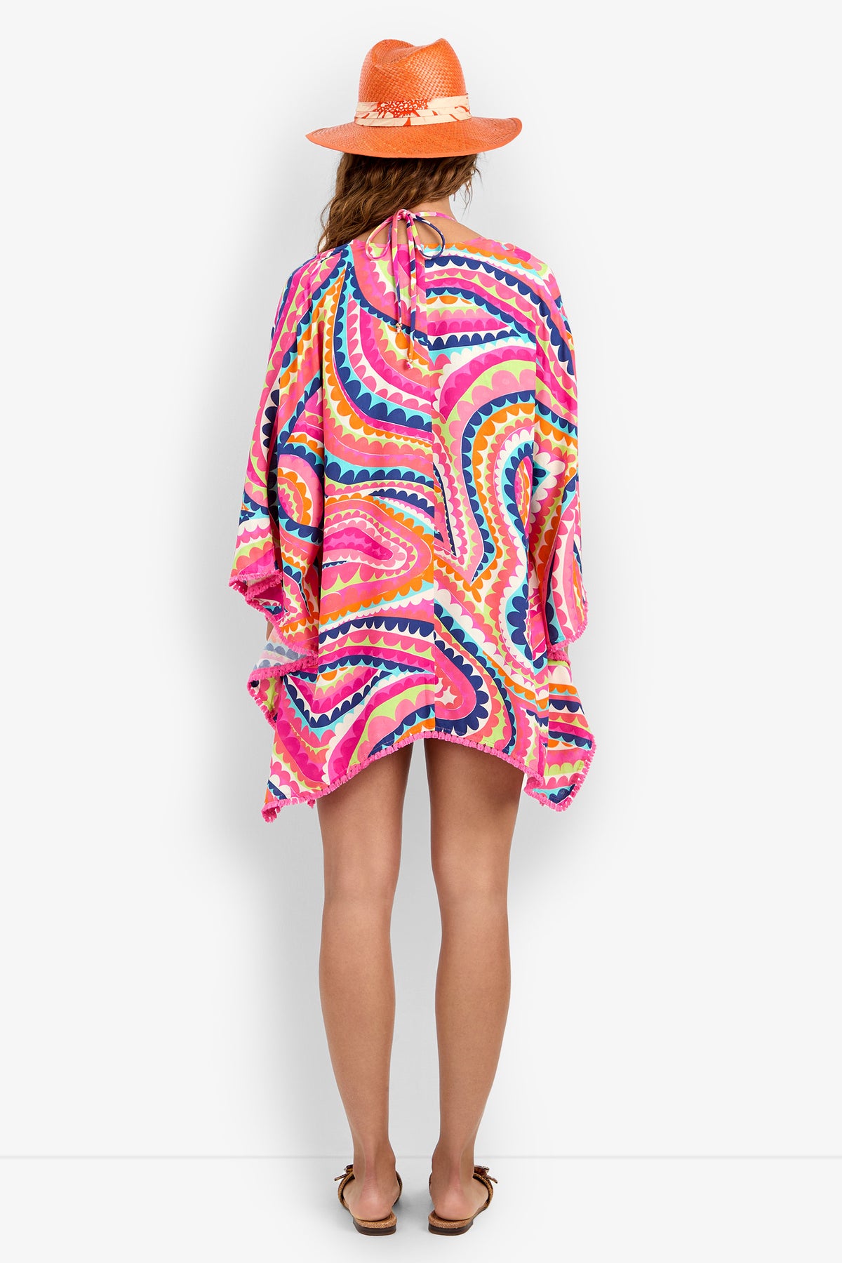 A person faces away, barefoot, wearing a bright orange hat and the Kimono Swim Cover-up—a loose piece featuring pink, orange, yellow, and blue swirling patterns that reaches mid-thigh.