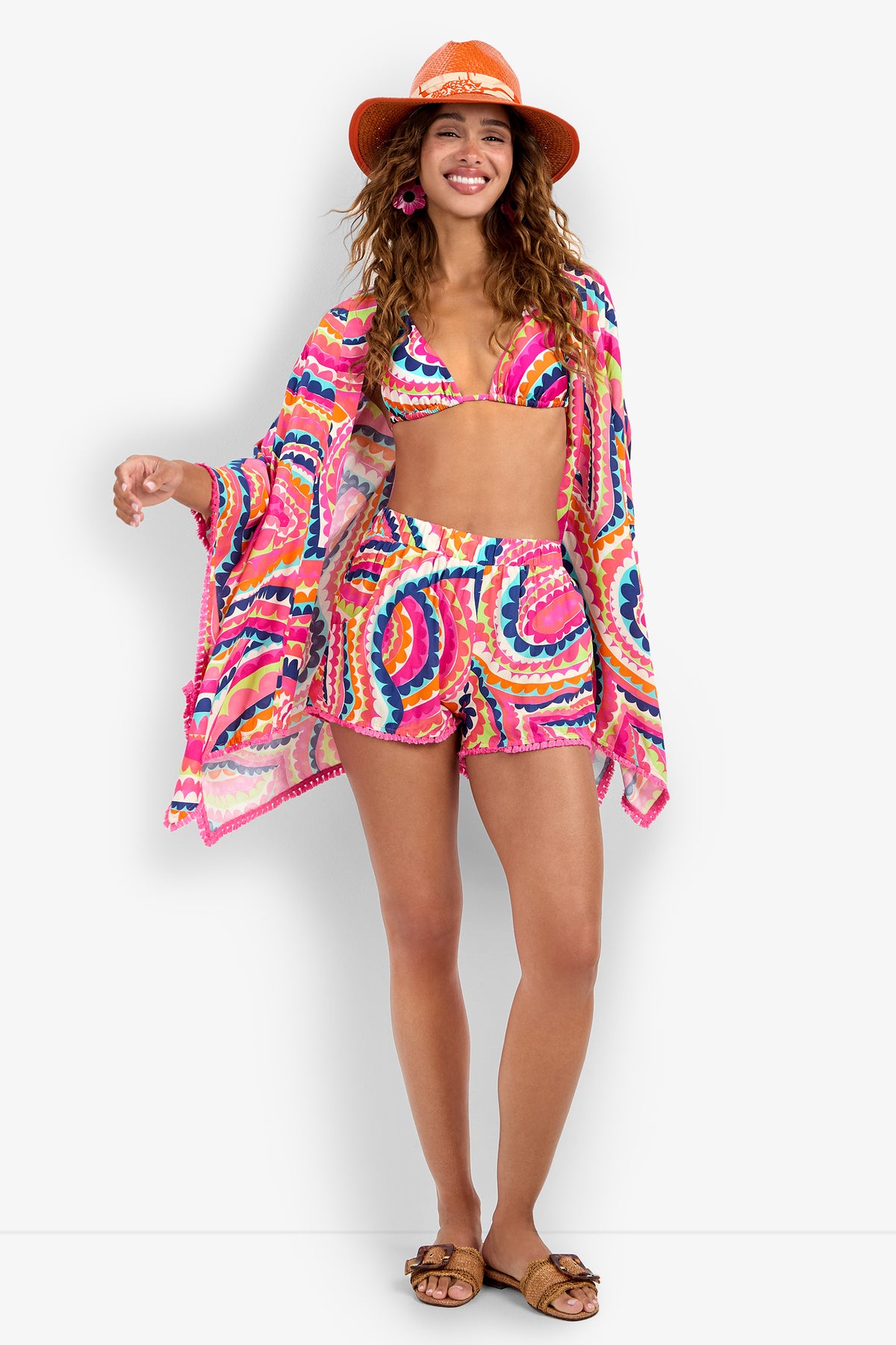 A woman smiles and poses in Aphrodite Swim Cover-Up Shorts with a matching bikini and cover-up, styled with an orange sunhat, pink earrings, and brown sandals against a white backdrop.