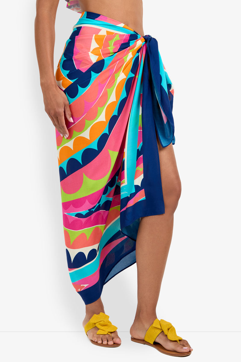A person wears the Pareo, a colorful patterned sarong with a navy border, tied at the waist and paired with yellow knotted sandals. Only the lower body is shown against a plain white background.