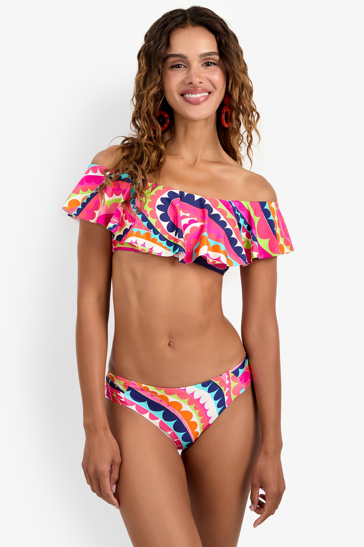 A woman smiles against a light background, wearing the Bandeau Bikini Top—an off-the-shoulder style with ruffles and bold wavy patterns in pink, orange, white, blue, and purple. She has long wavy hair.