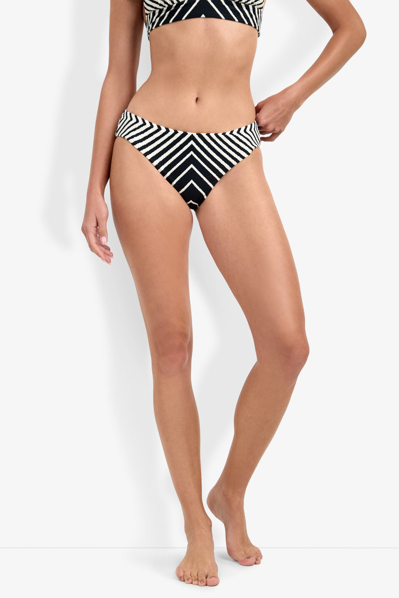 A woman stands barefoot against a plain white background, wearing the Net Stripe Hipster Bikini Bottom in black with white geometric stripe patterns; only her body from shoulders to feet is visible.