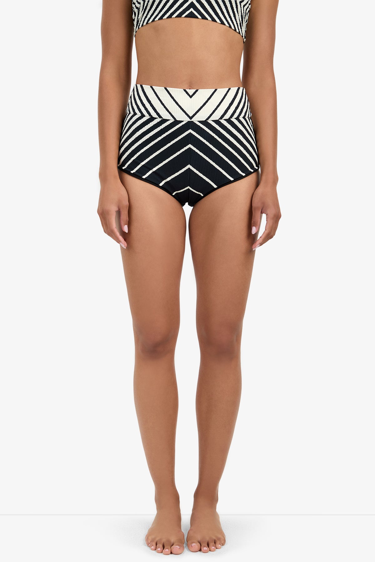 A woman stands barefoot in Net Stripe Swim Shorts, featuring bold black and white high-waisted chevron stripes. The image is cropped from shoulders to feet, set against a plain white background.