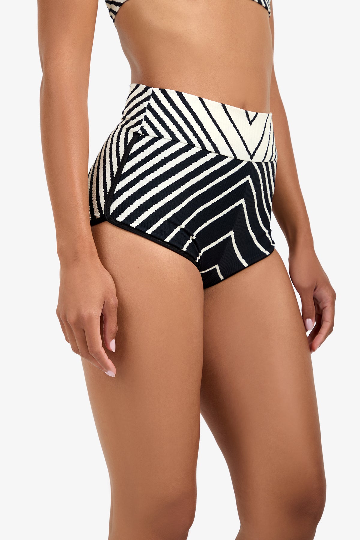 Wearing the Net Stripe Swim Short, a woman poses against a plain white background. The high-waisted design features bold black and white geometric stripes.