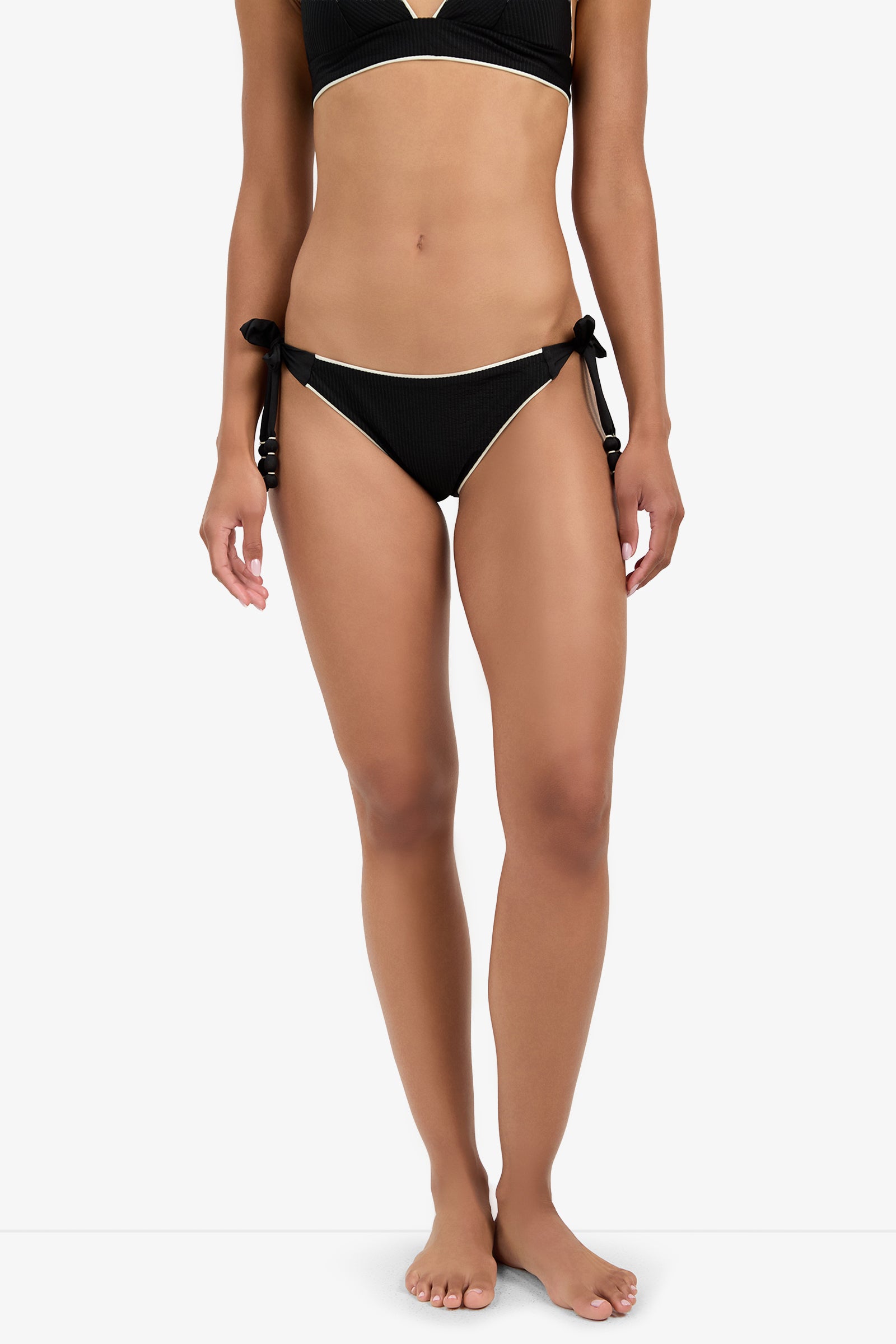 A woman stands barefoot wearing the CORINTH TIE SIDE HIPSTER bikini, black with white trim and side-tie bottoms, shown from shoulders to feet against a plain white background.