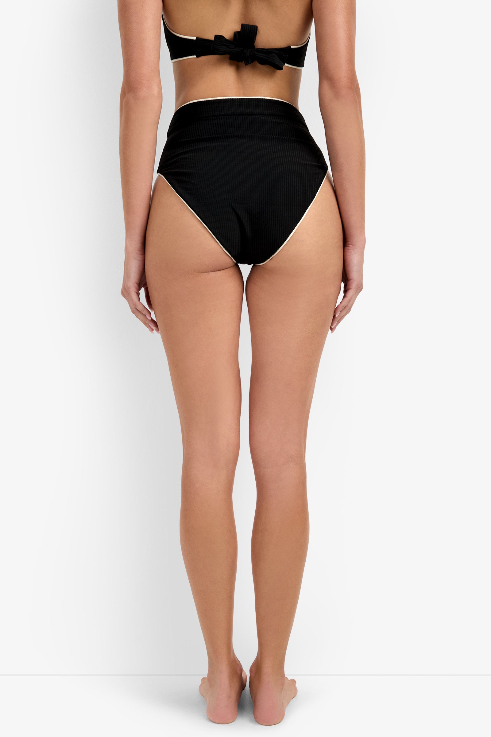 A woman stands barefoot, facing away from the camera, wearing the Corinth High Waist Bikini Bottom and a matching black bikini top with a bow at the back against a plain white background.