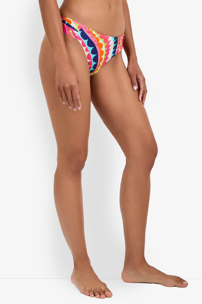 A woman stands barefoot against a plain white background, wearing the Shirred Hipster bikini bottoms featuring a bold, wavy pattern in pink, orange, blue, and white.