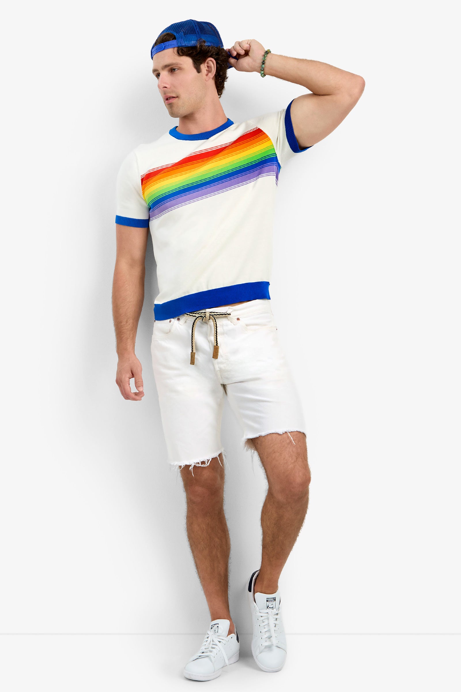 A man wearing the Staley Pride Ringer—a white T-shirt with a rainbow stripe—along with a blue cap, frayed white shorts with a rope belt, and white sneakers stands against a white background, looking to the side.