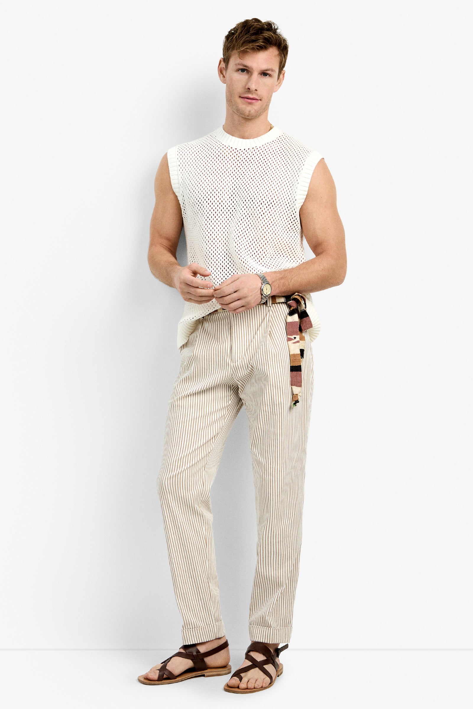 A man stands against a white background wearing the KRAMER MESH PULLOVER, paired with light striped pants, a patterned belt, brown sandals, and a wristwatch. He gazes at the camera with a relaxed expression.