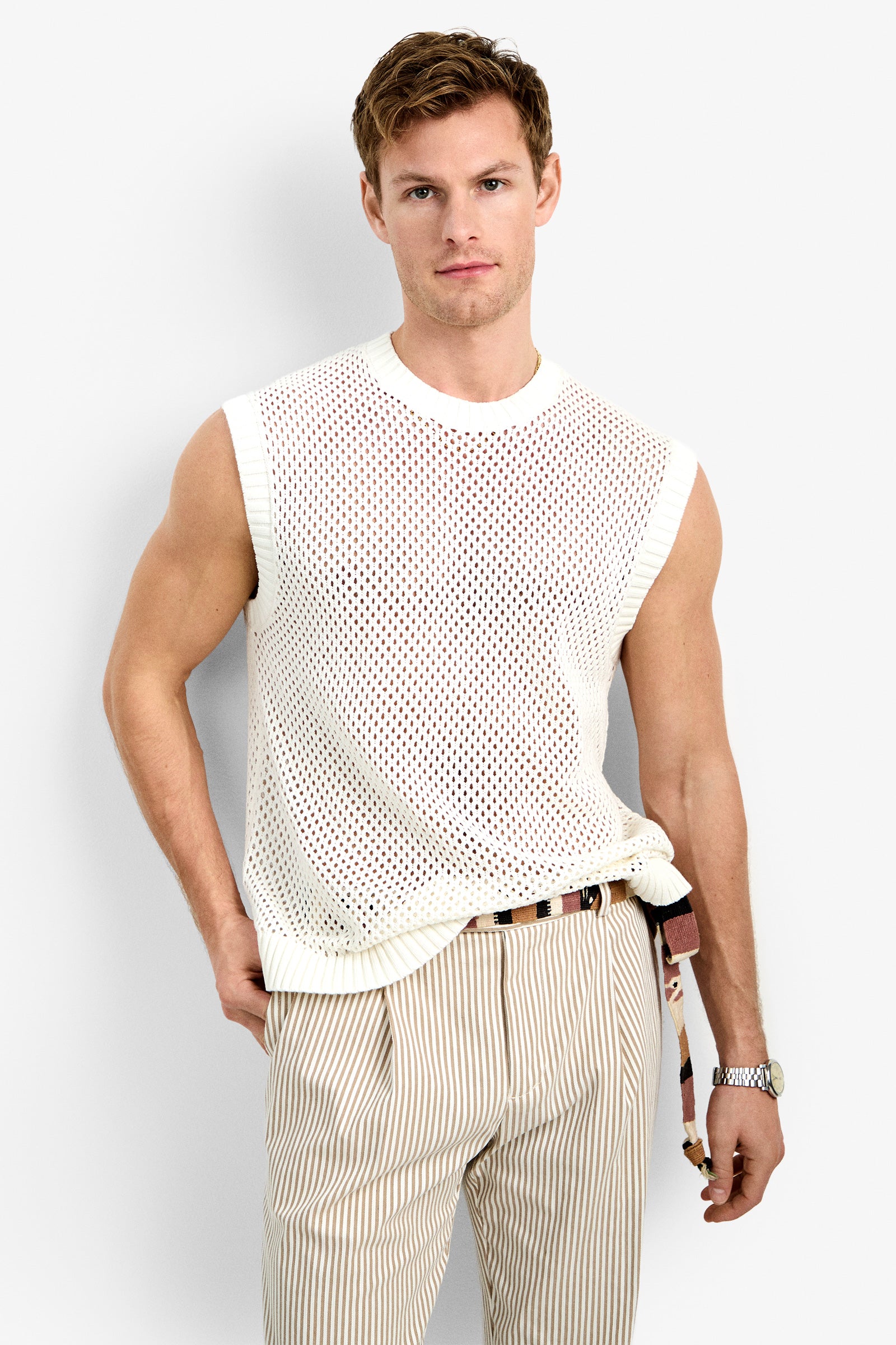 A man wears the KRAMER MESH PULLOVER, a sleeveless white knit mesh top, paired with beige striped pants and a belt, standing against a plain light background and looking at the camera with a neutral expression.