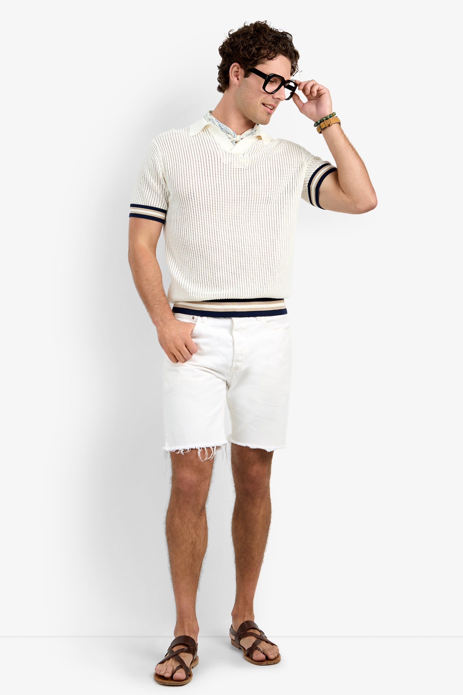 A man wearing black glasses, the Alvin Mesh Polo over a collared shirt, white frayed shorts, brown sandals, and bracelets stands smiling, adjusting his glasses.