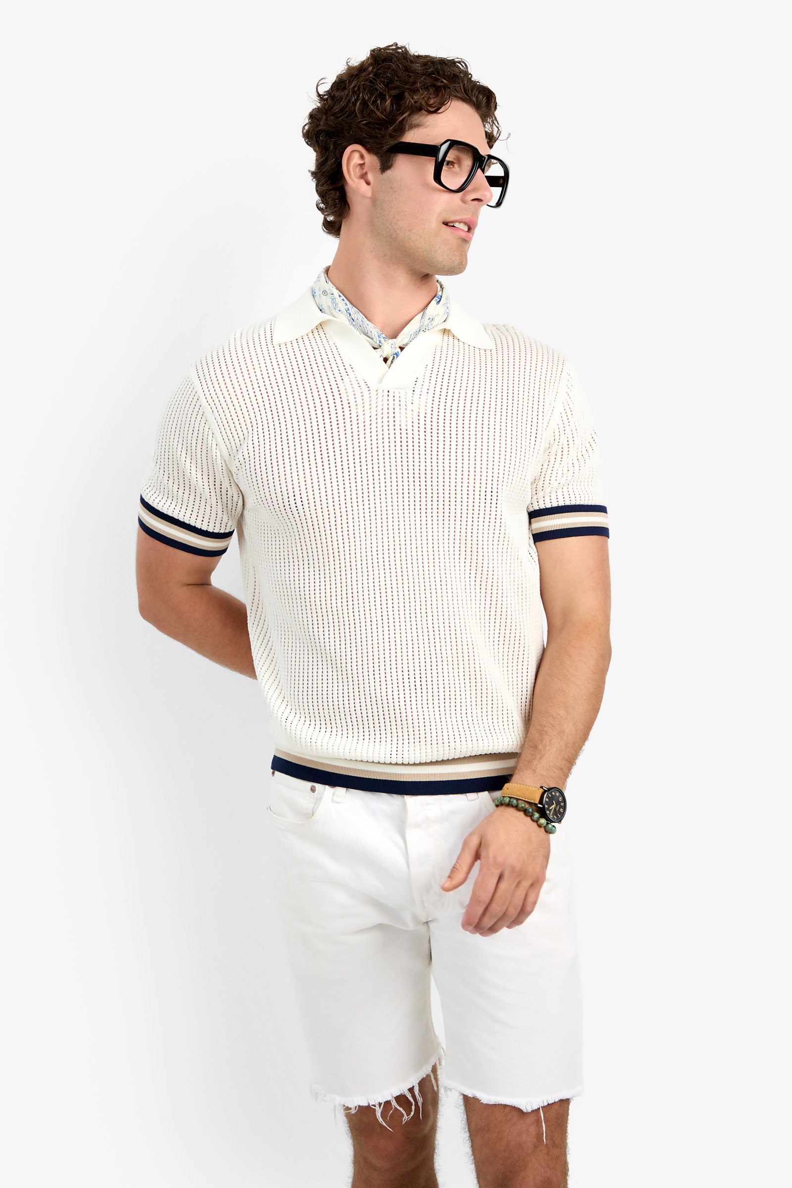 A man with curly hair and black glasses wears the Alvin Mesh Polo—a cream short-sleeve sweater with navy trim—over a collared shirt, paired with white shorts and a black watch, standing against a plain white background.