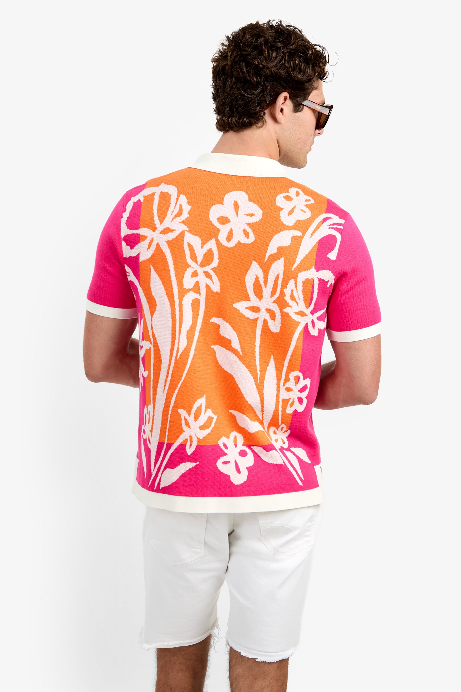 A man faces away, showcasing the Alessio Polo—a vibrant pink and orange shirt with bold white floral patterns, paired with sunglasses and white shorts.