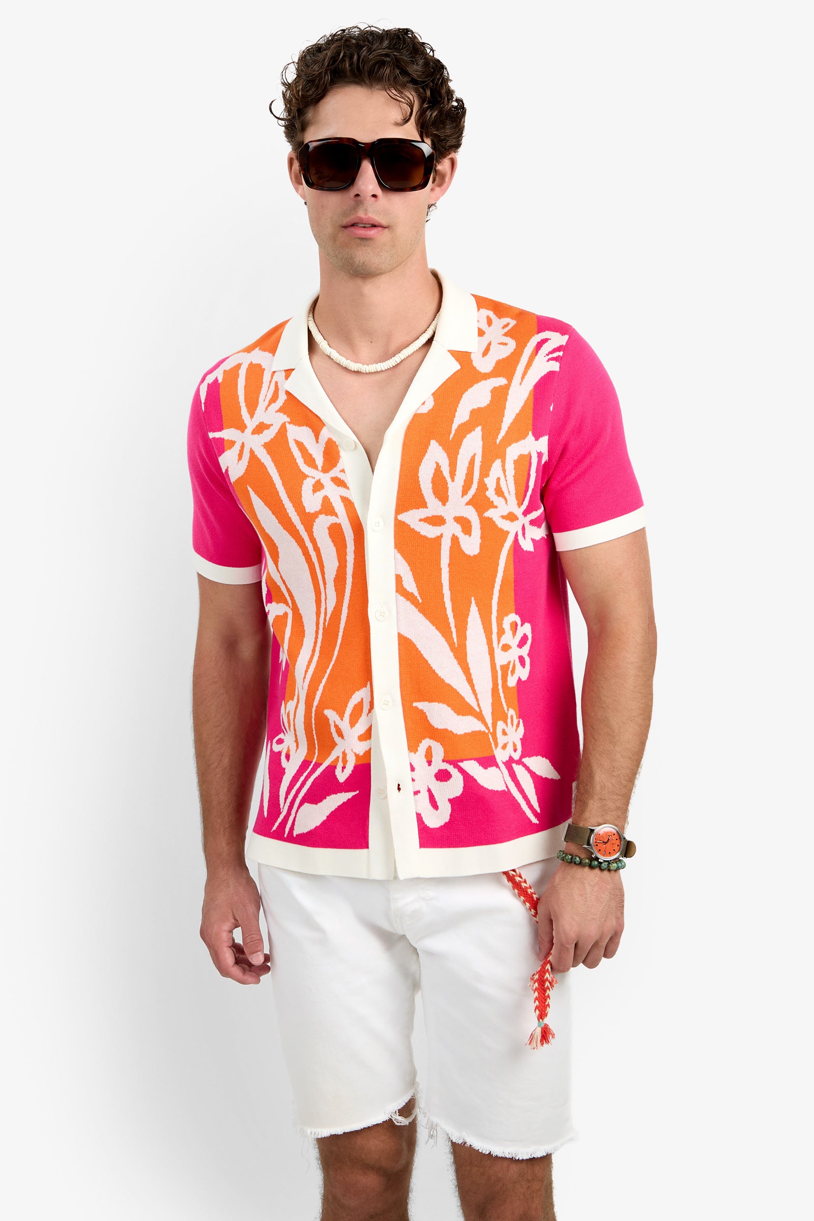A man wearing dark sunglasses, the Alessio Polo—a colorful short-sleeve shirt with a pink, orange, and white floral pattern—white frayed shorts, a beaded necklace, and bracelets stands against a plain white background.