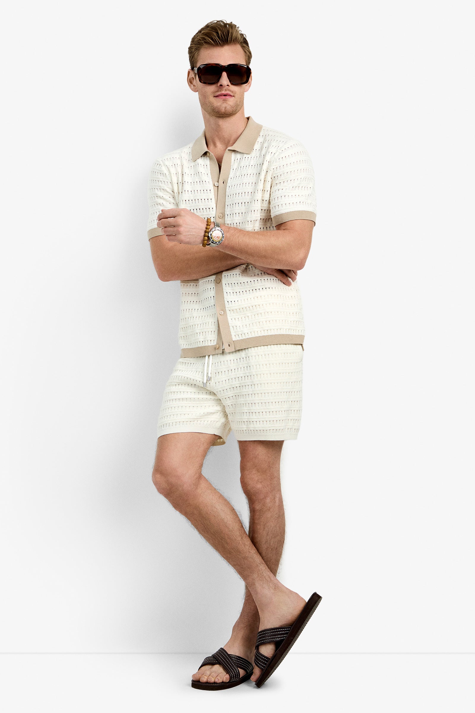 A man in sunglasses, a watch, and dark slide sandals stands with legs crossed against a plain white background, wearing the Hillcrest Resort Polo—a matching cream-colored 100% cotton knit short-sleeve shirt and shorts.