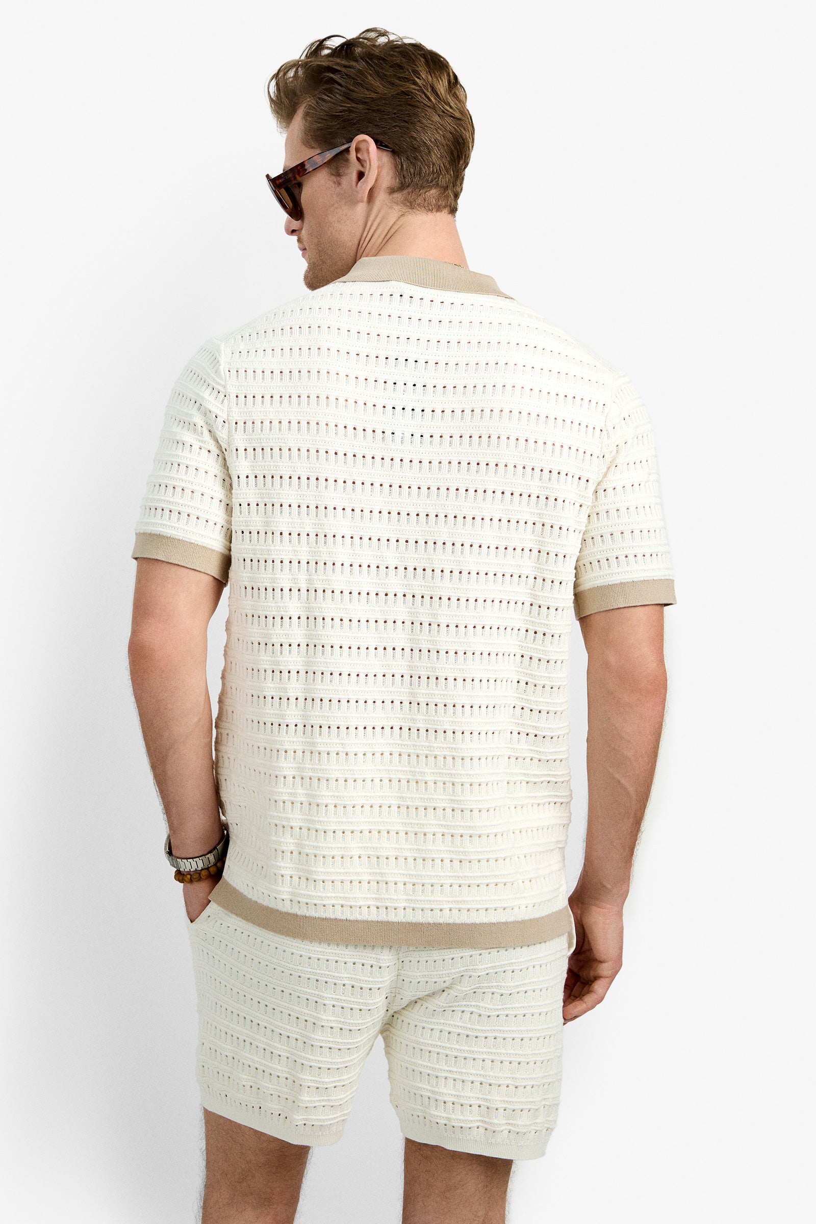A man with light brown hair, wearing sunglasses and the Hillcrest Resort Polo—a cream-colored, 100% cotton crochet short-sleeve shirt—stands with his back to the camera against a plain white background.