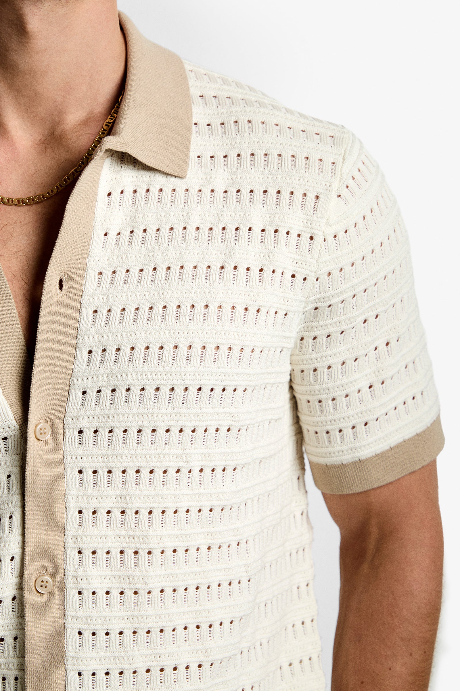 A person wears the Hillcrest Resort Polo, a cream-colored, soft cotton knit with short sleeves, beige collar and cuffs, and a textured perforated pattern, paired with a gold chain necklace.