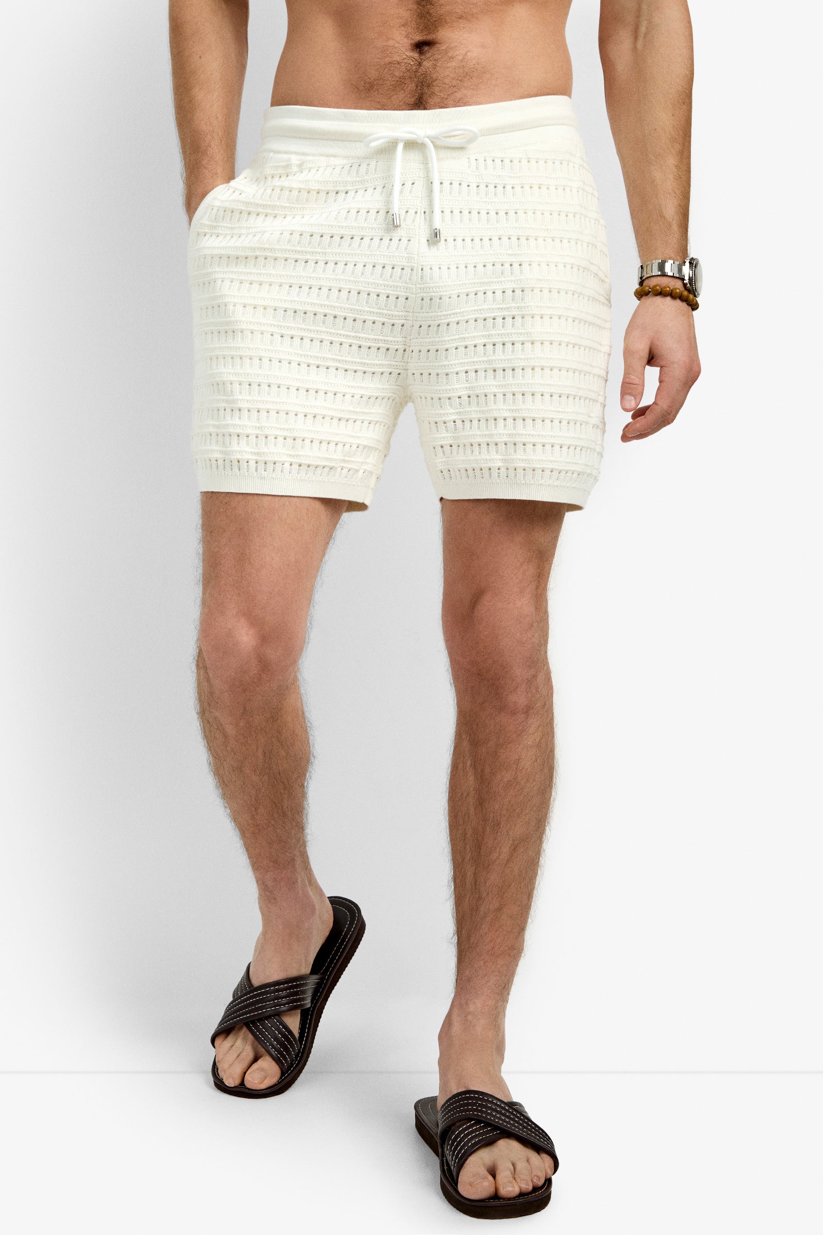 A man stands against a plain white background wearing the Julio Crochet Short in cream—a textured, 100% cotton knit with a drawstring waist—paired with black sandals and a watch.