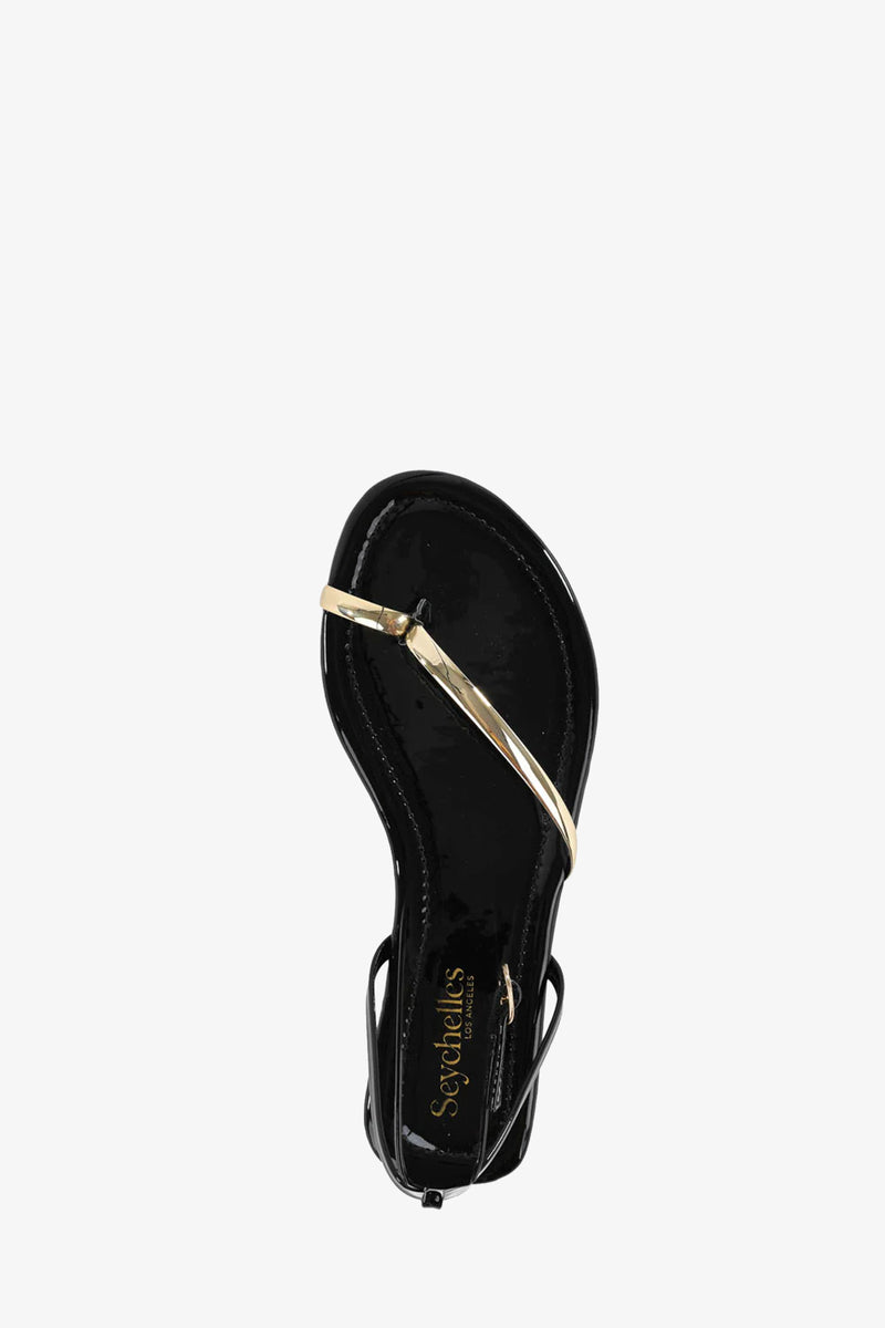 The A Little Extra Sandal is a black sandal with a shiny gold crisscross strap over the toe area, seen from above, and features the Seychelles brand name printed on the insole.