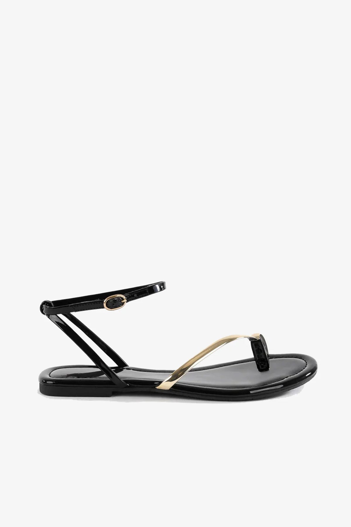 The A Little Extra Sandal, a black flat sandal featuring a thin ankle strap with a gold buckle and a gold accent on the toe strap, is displayed on a plain white background.
