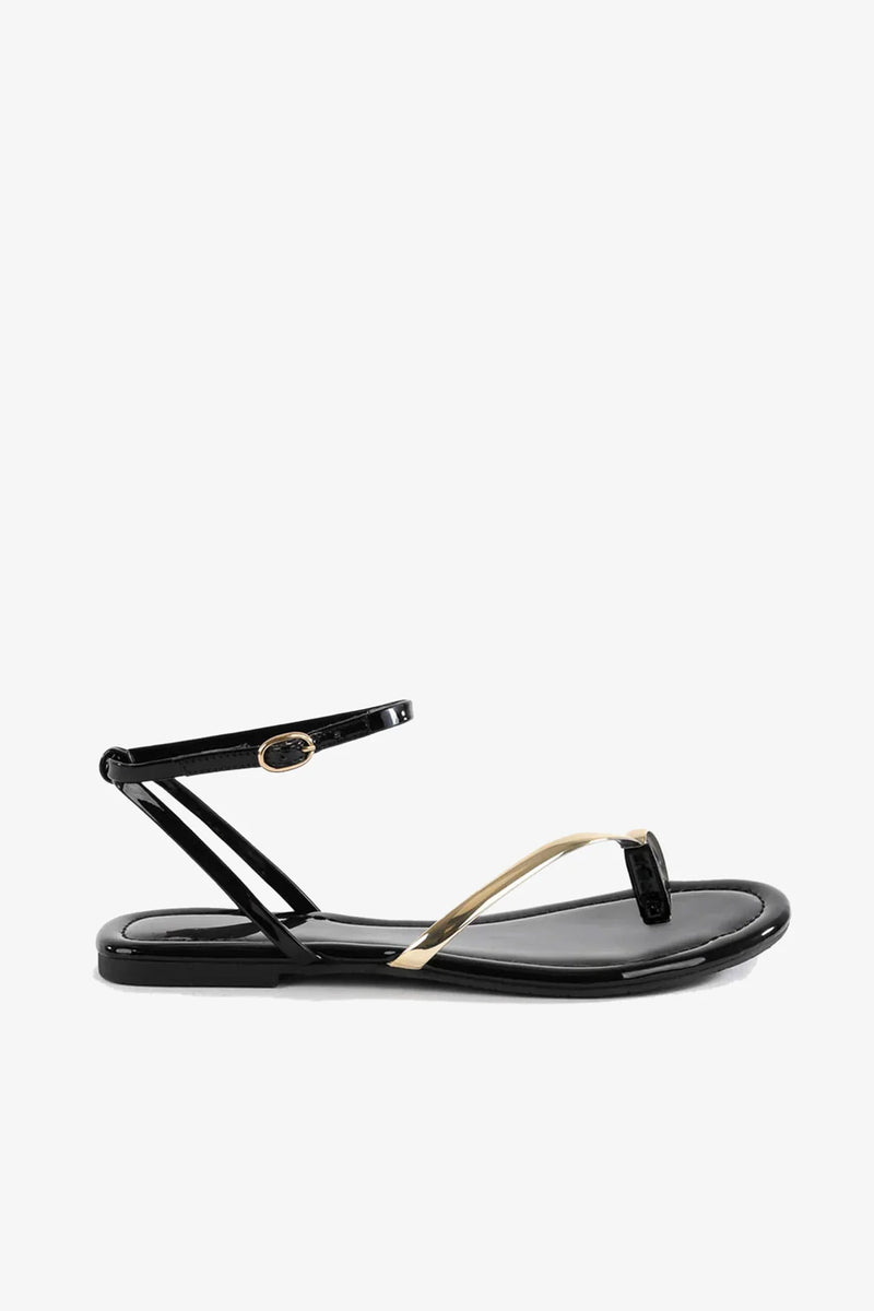 The A Little Extra Sandal, a black flat sandal featuring a thin ankle strap with a gold buckle and a gold accent on the toe strap, is displayed on a plain white background.