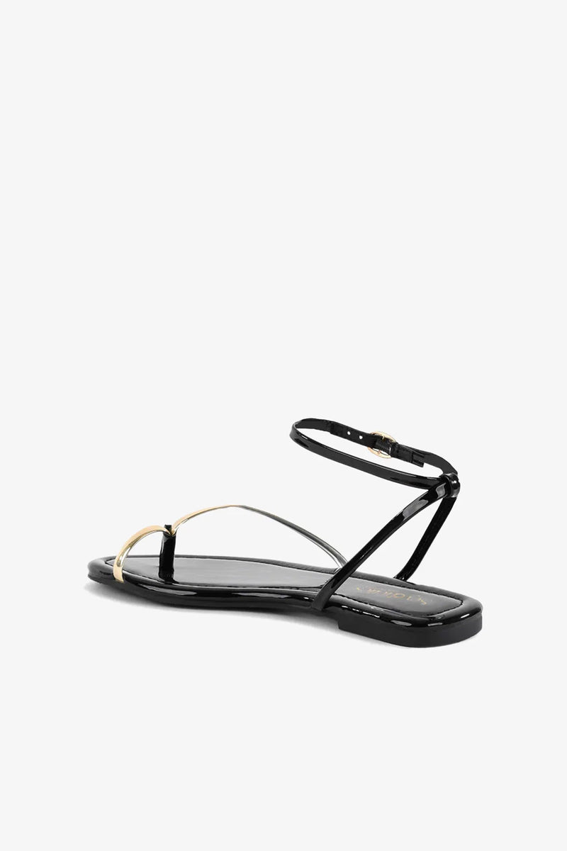 A Little Extra Sandal in black features a thin ankle strap and a metallic gold accent across the toes, shown against a plain white background.