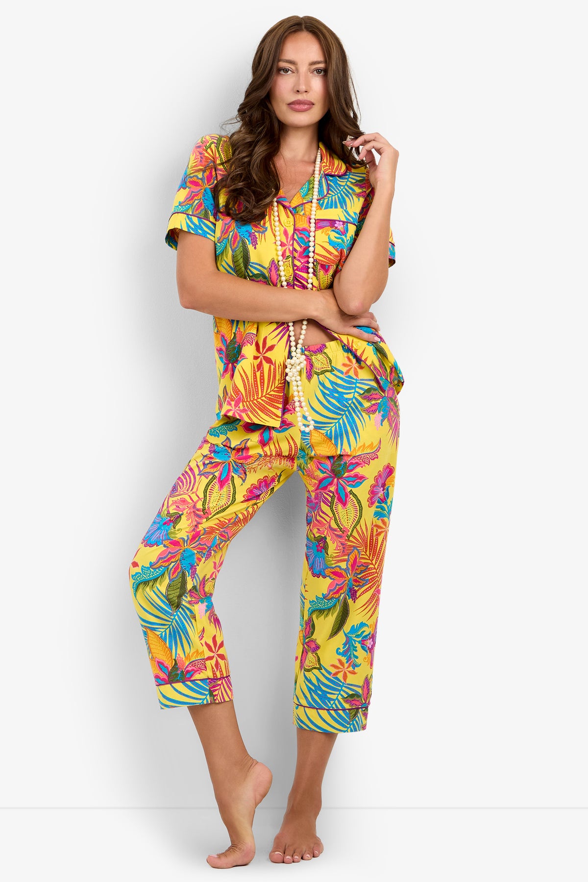 A woman wearing the Jacobean Short Sleeve Cropped PJ Set, featuring a vibrant yellow hue with colorful tropical floral patterns, stands barefoot against a white background—one hand gently touching her face and the other holding a beaded necklace.