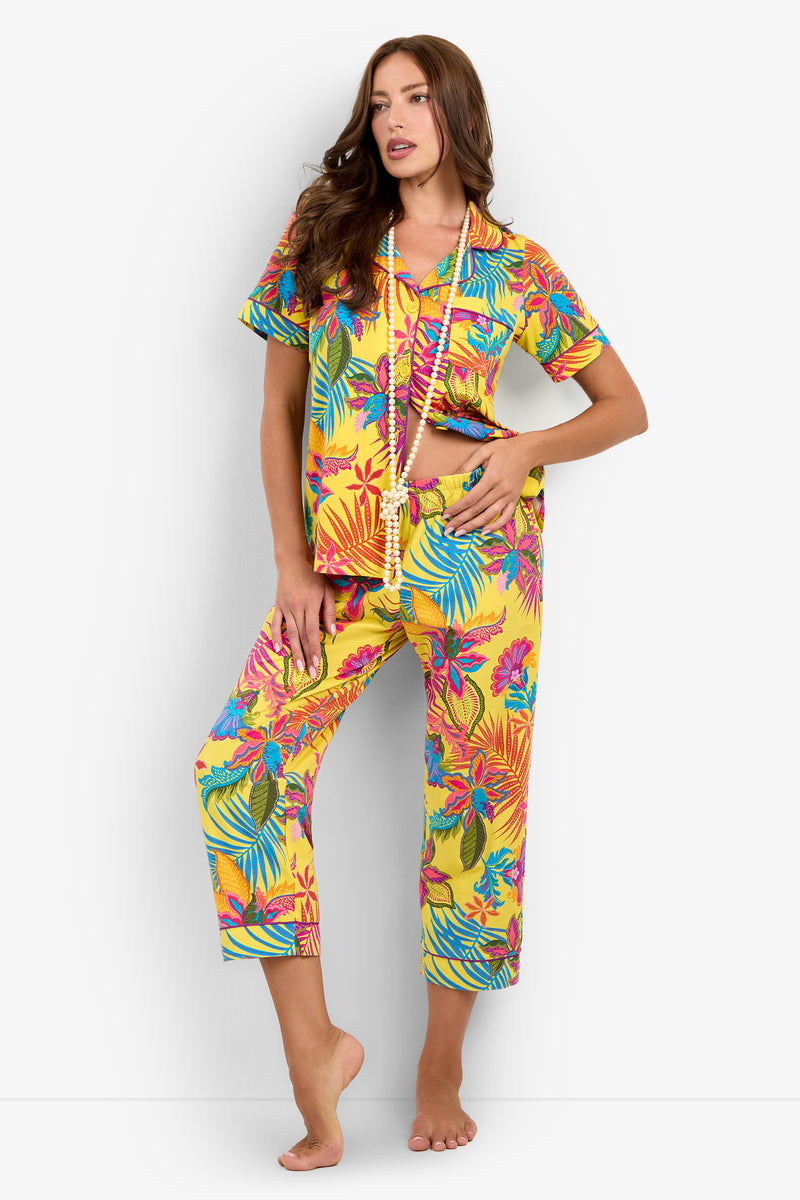 A woman stands barefoot against a plain white background, wearing the Jacobean Short Sleeve Cropped PJ Set in a vibrant yellow tropical print and accessorized with two long beaded necklaces.