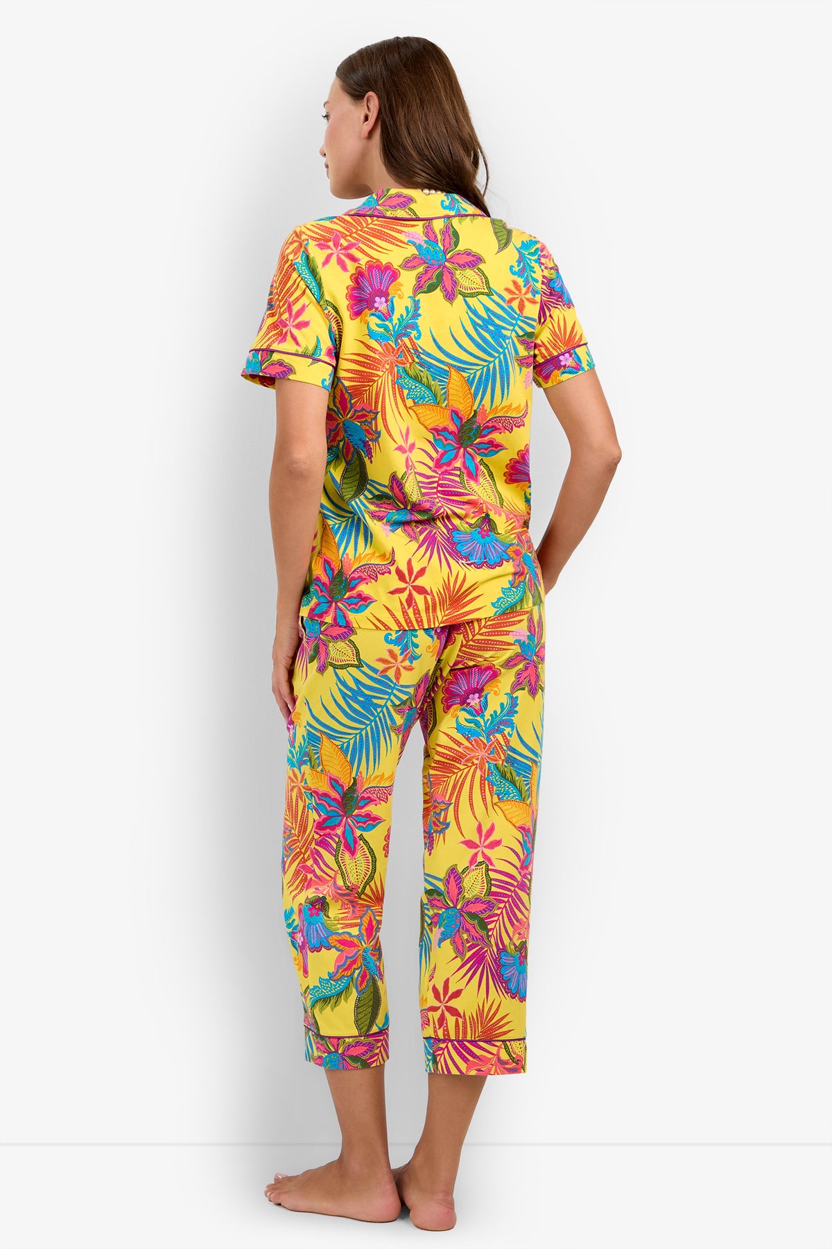 A woman stands barefoot, facing away, wearing the Jacobean Short Sleeve Cropped PJ Set featuring bright yellow fabric adorned with a colorful floral and leaf print in pink, blue, and green. The pajama set includes short sleeves and cropped pants.