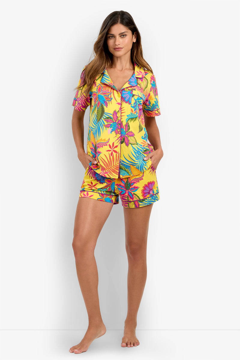 A woman stands barefoot against a plain white background wearing the Jacobean Short Sleeve PJ Set, featuring a yellow base adorned with colorful tropical floral patterns, including blue, pink, and green leaves and flowers.