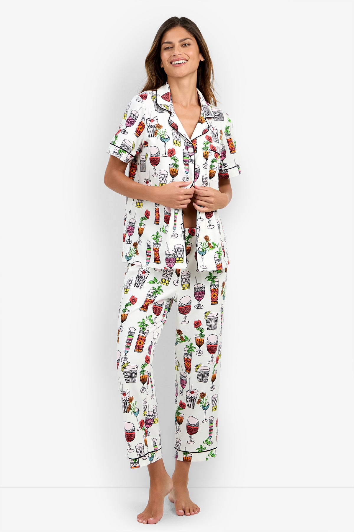 A woman stands and smiles, wearing the Short Sleeve Cropped Pajamas Set featuring a white base with colorful drink and cocktail glass prints, a short-sleeve button-up top, and matching long pants. The photo is taken against a plain, light-colored studio background.
